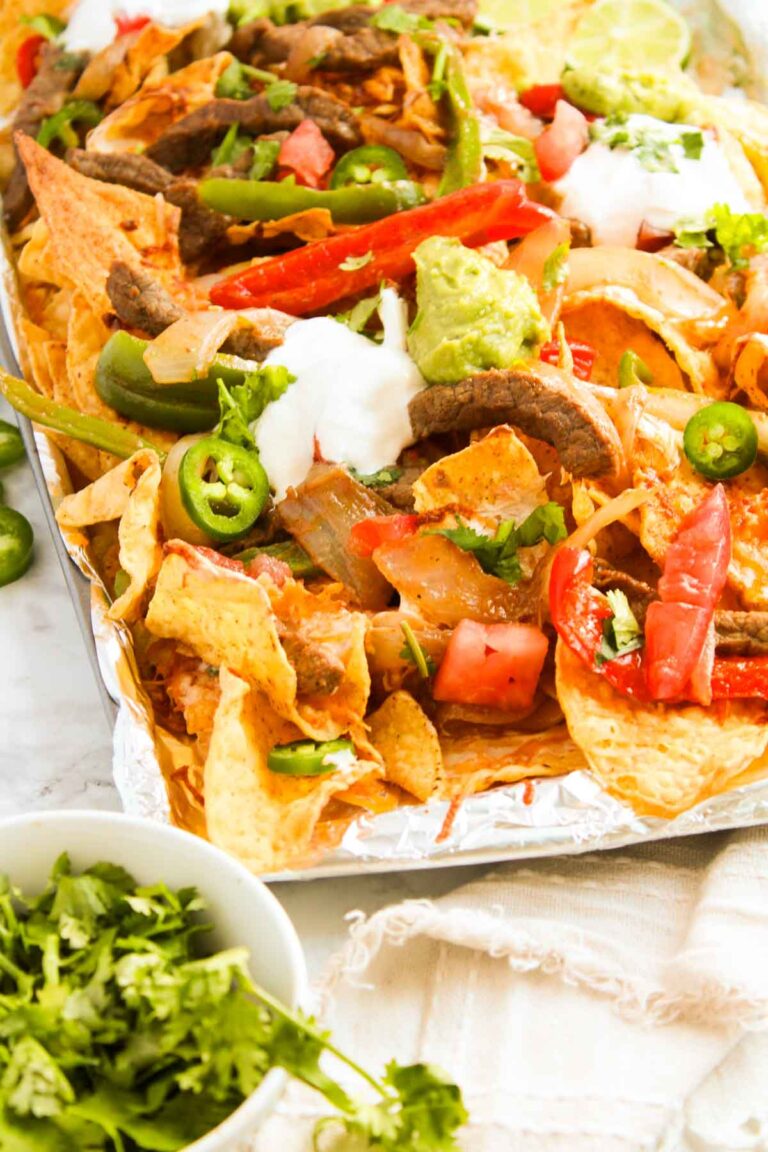 Close-up of steak fajita nachos topped with jalapeños, tomatoes, onions, guacamole, sour cream, and cilantro, all served on a foil-lined tray.