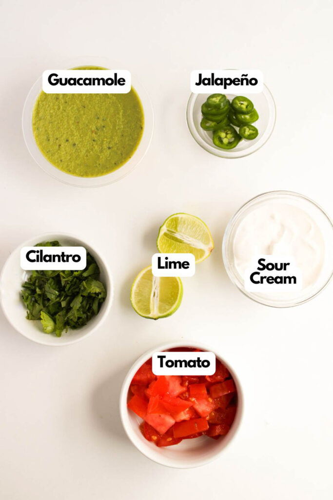 Five bowls containing guacamole, jalapeño slices, sour cream, cilantro, and diced tomato are arranged around lime wedges on a white surface, creating the perfect accompaniments for a platter of steak fajita nachos.