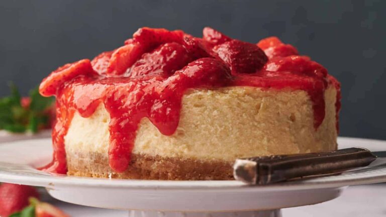 Cheesecake topped with fresh strawberry sauce on a white cake stand, with a cake server beside it.
