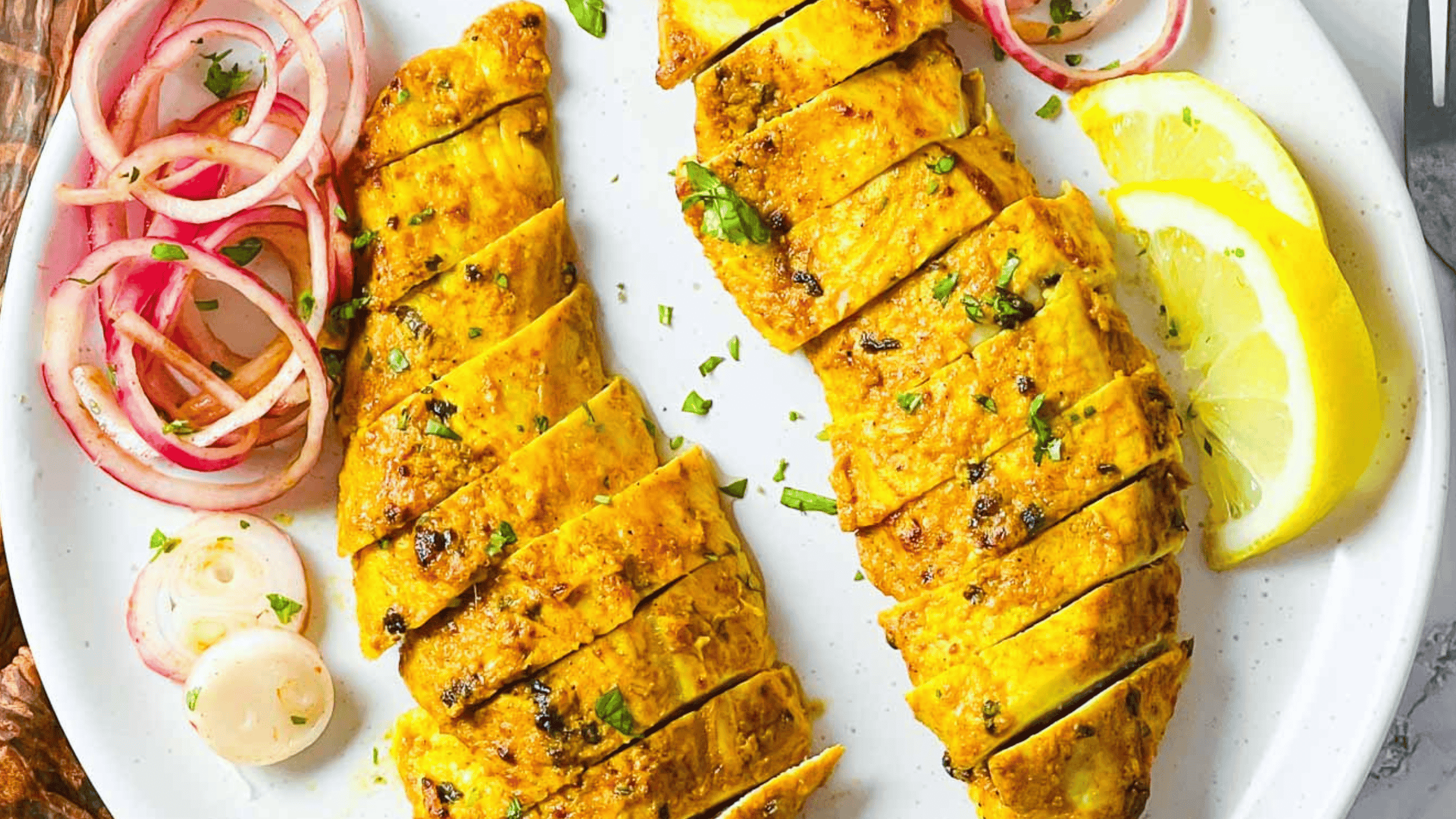 Two Tandoori Turkey steak with herbs, lemon slices, and pickled onions on a white plate.