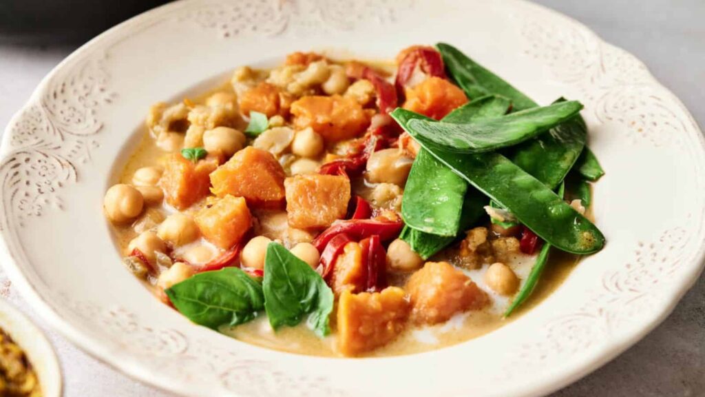 A white bowl with a stew containing sweet potatoes, chickpeas, tomatoes, and green snow peas, garnished with spinach leaves.