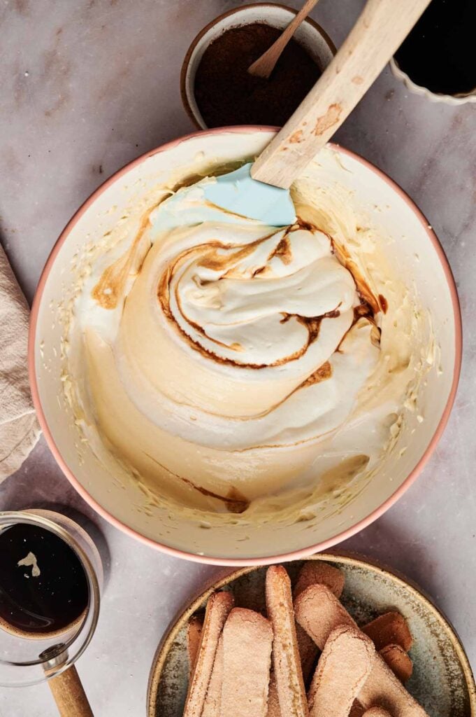 A mixing bowl brimming with creamy tiramisu filling is being stirred gently with a spatula, while ladyfingers and other essential ingredients await their turn to create the perfect dessert.