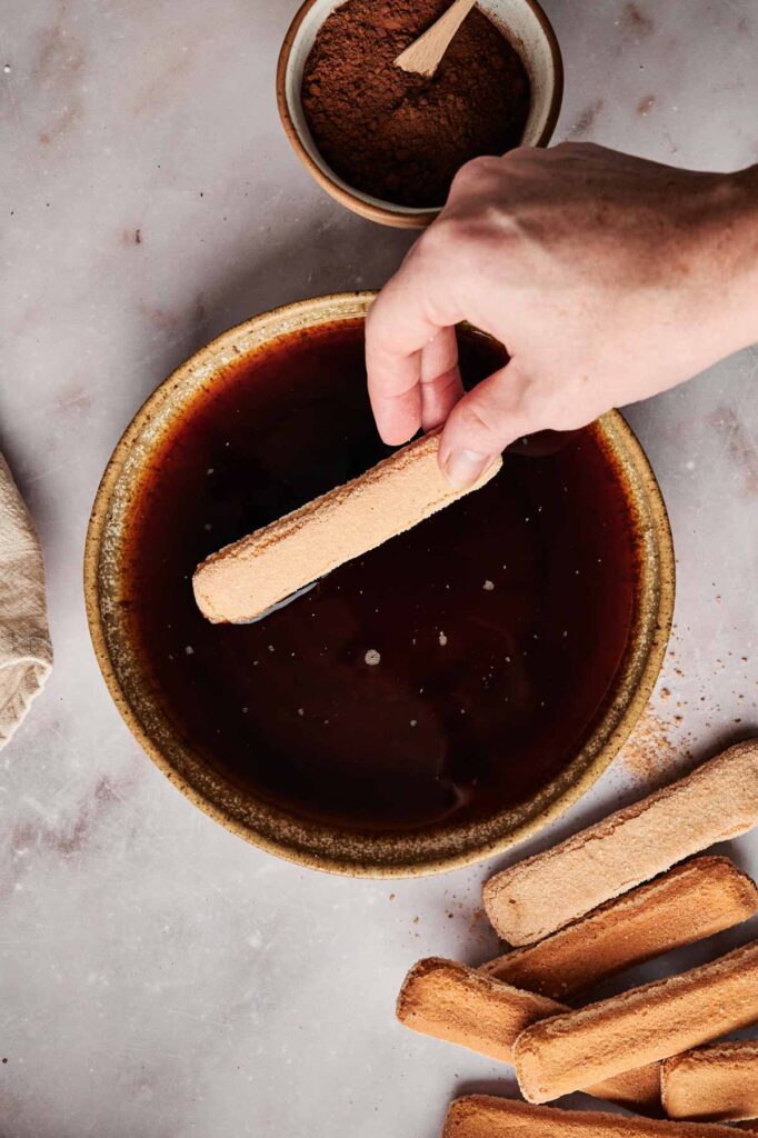 A person's hand dips a ladyfinger biscuit into a bowl of coffee, setting the stage for tiramisu. A small bowl of cocoa powder and more ladyfingers sit nearby on the marble surface.