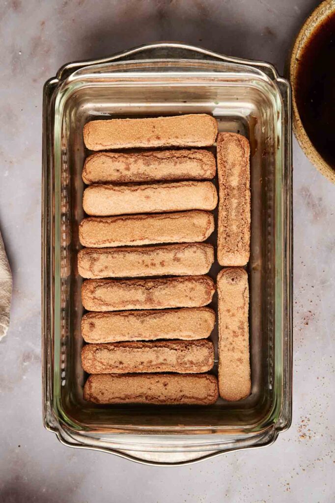 A glass dish showcasing a single layer of ladyfinger cookies, dusted with cocoa powder, evokes the essence of classic tiramisu on a gray surface.
