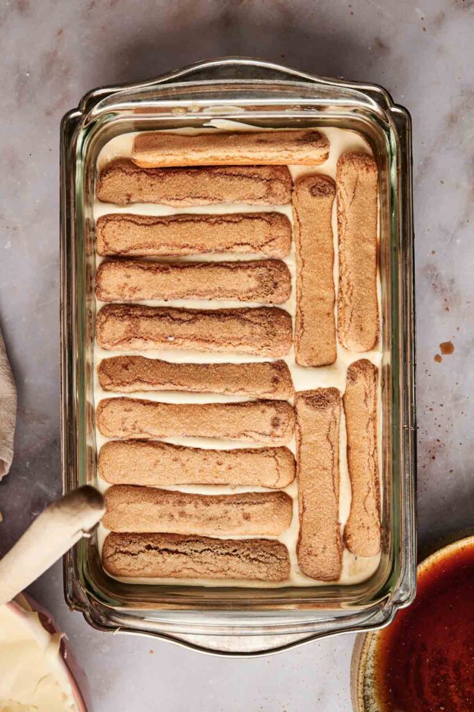 A glass dish with layered tiramisu showcases elegant layers of cream filling and soaked ladyfingers. Beside it, a mixing utensil rests, while a partial view of a bowl with liquid adds to the atmosphere.