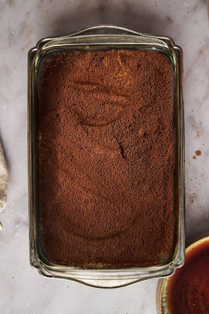 A glass baking dish brimming with creamy tiramisu rests elegantly on a marble surface, its top adorned with a delicate dusting of cocoa powder.
