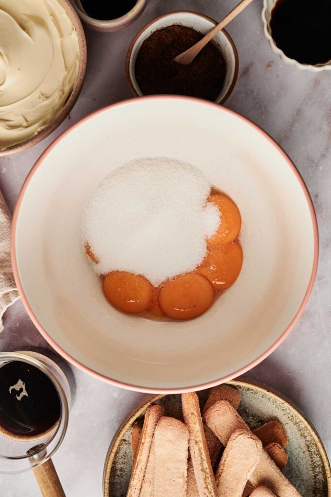 Egg yolks and sugar blend seamlessly in a mixing bowl, surrounded by essential tiramisu ingredients like mascarpone, coffee, and ladyfinger biscuits, ready to create the classic dessert.