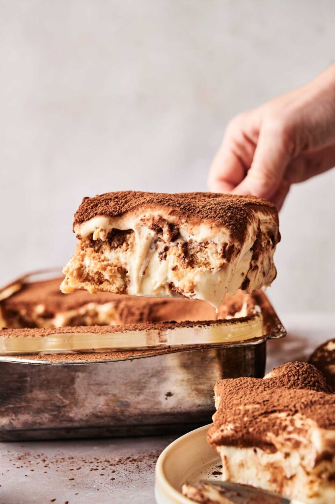 A hand delicately lifts a slice of tiramisu from the rectangular dish, revealing its luscious layers of cream and cocoa. A portion is elegantly served on a plate below, ready to delight.