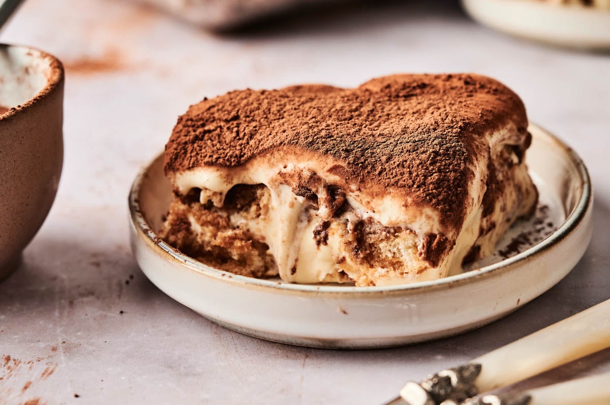 A slice of indulgent tiramisu rests on a round plate, its cocoa-dusted surface inviting. Two forks lie nearby on the table, ready to share this delightful dessert experience.