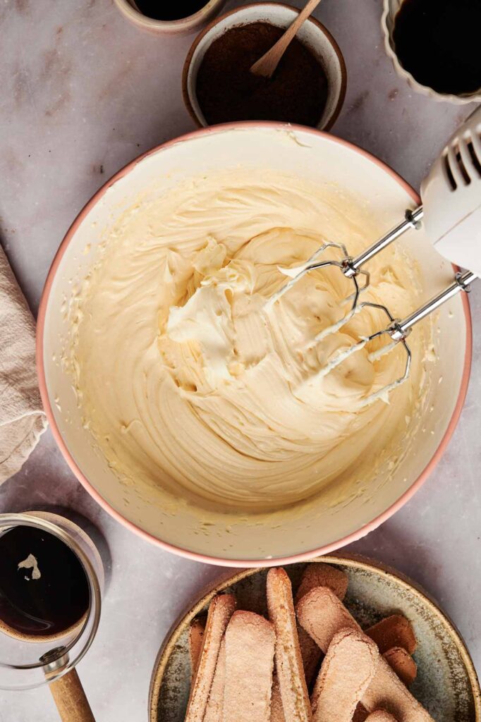 A bowl of creamy tiramisu mixture being blended with a hand mixer, surrounded by coffee cups, a cloth, and ladyfingers on the table.