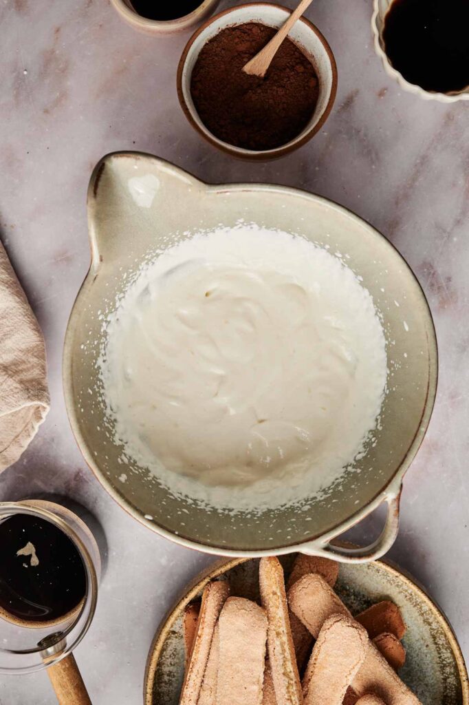 A mixing bowl filled with a creamy white tiramisu mixture, surrounded by bowls of cocoa powder, coffee, and ladyfinger cookies on a marble surface.