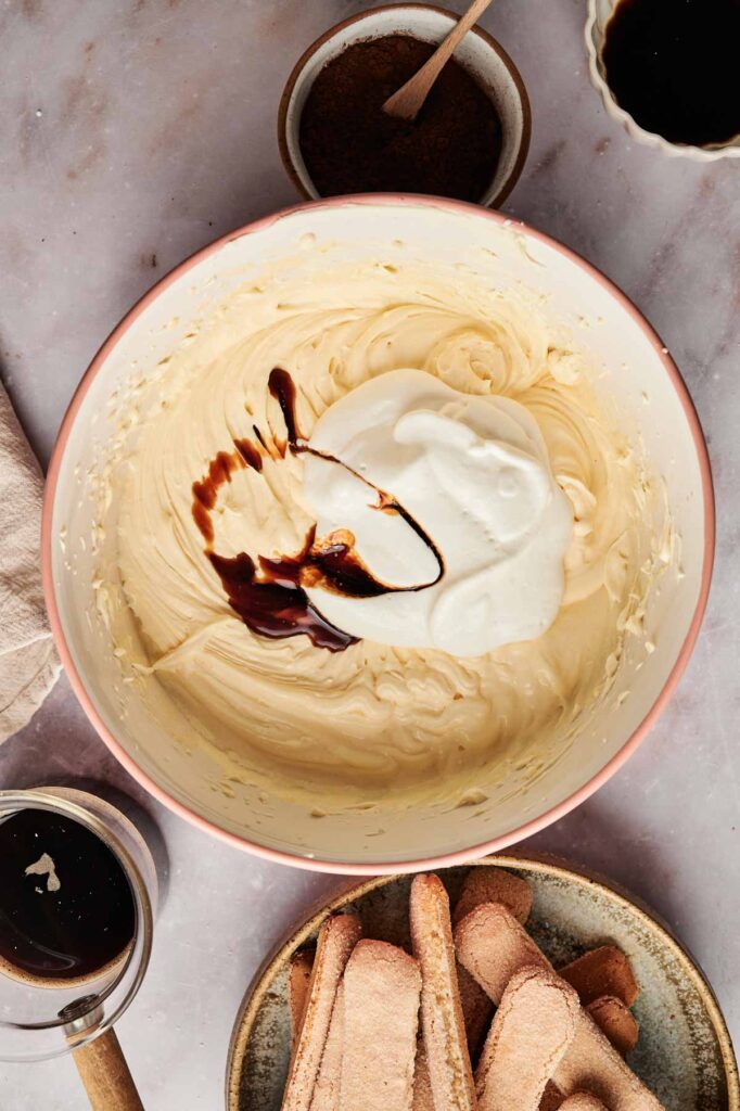 A mixing bowl brimming with a creamy tiramisu mixture, blending white cream and dark liquid. Surrounding bowls hold coffee and ingredients, while savoiardi biscuits rest on a nearby plate.