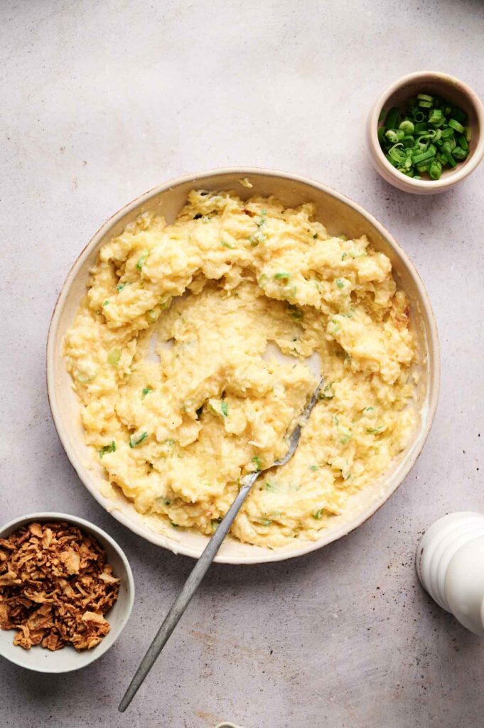 A bowl of twice-baked mashed potatoes with a spoon invites indulgence. Surrounding it, bowls of green onions and crispy onions rest on a light surface, ready to enhance the dish.