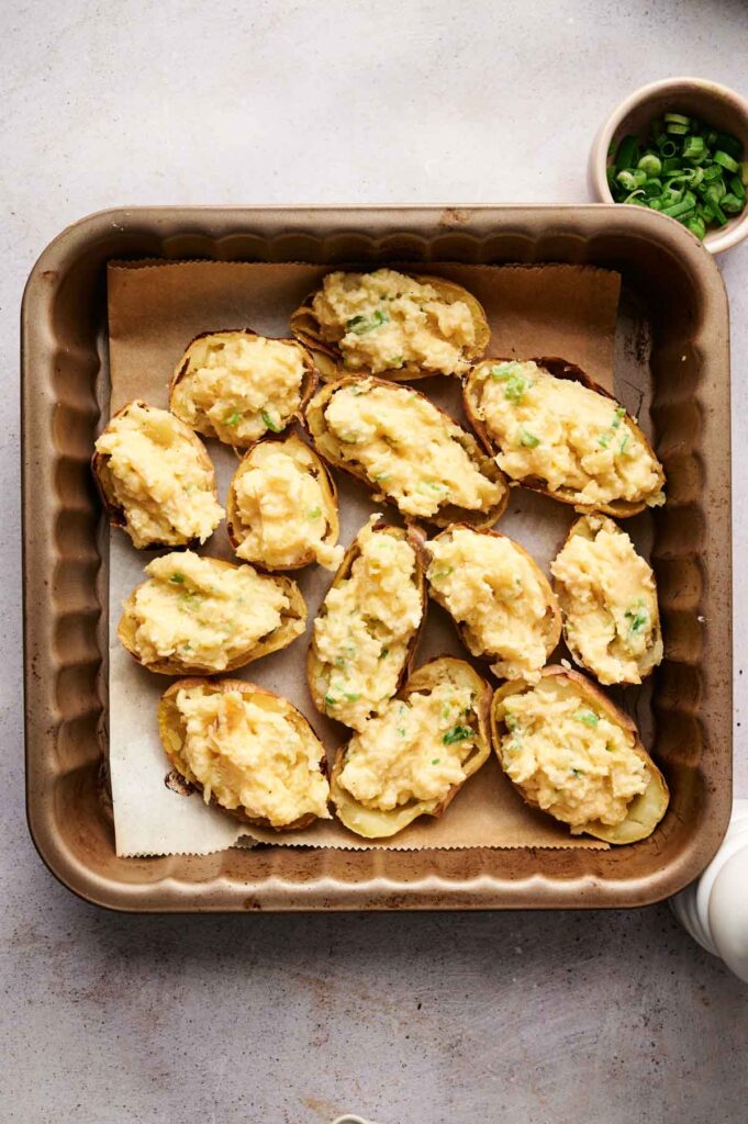 Twice-baked potato skins filled with a creamy, cheesy mixture and topped with green onions are perfectly arranged in a baking dish.