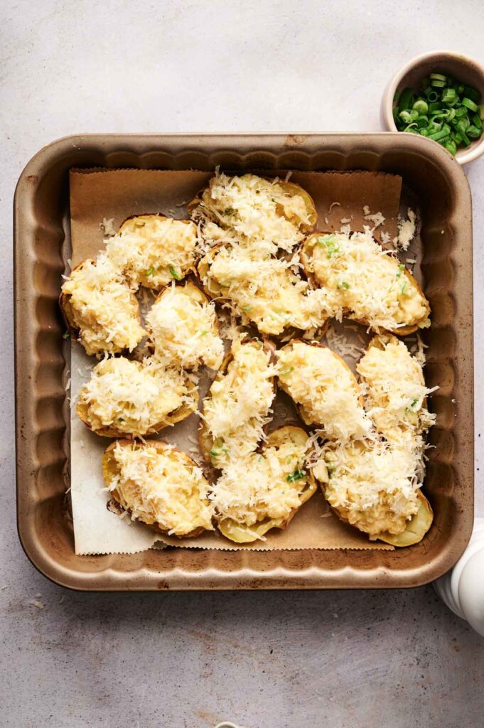 Twice-baked potato skins topped with cheese and chopped green onions in a square baking dish, with extra green onions in a small bowl beside it.