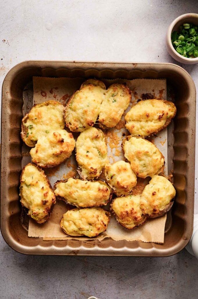 A baking tray holds twelve twice-baked potatoes topped with cheese, their golden crusts glistening. In the corner, a small bowl of chopped green onions adds a pop of color to these delicious treats.