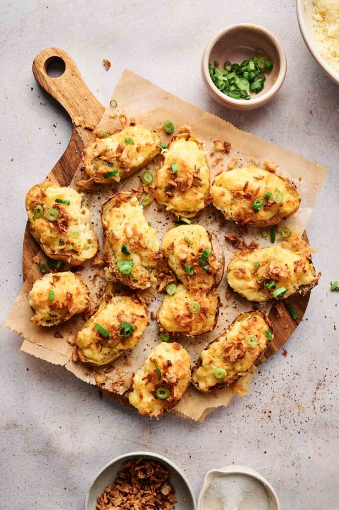 A wooden board showcases twice-baked potatoes crowned with melted cheese, chopped green onions, and crispy bits. Beside them are small bowls filled with extra green onions and a savory seasoning to enhance the flavor.