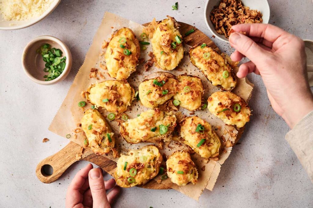 Hands arranging twice baked potato halves loaded with cheese and green onions on a wooden board.