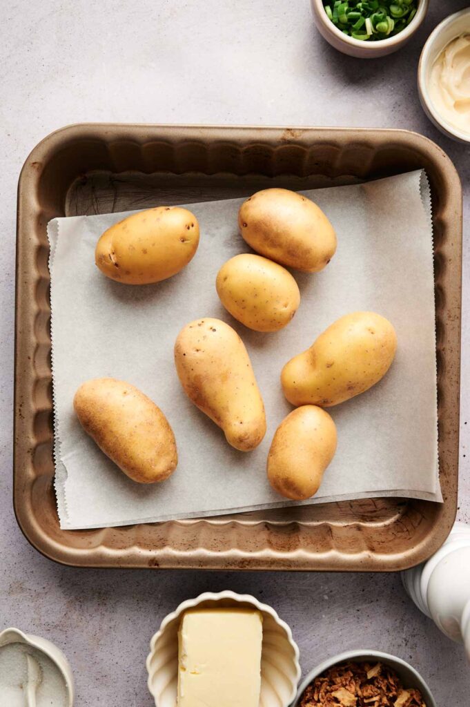 Seven potatoes rest on parchment paper in a baking tray, ready to become delicious twice baked potatoes. They're surrounded by butter, chopped green onions, and other tempting ingredients on the table.