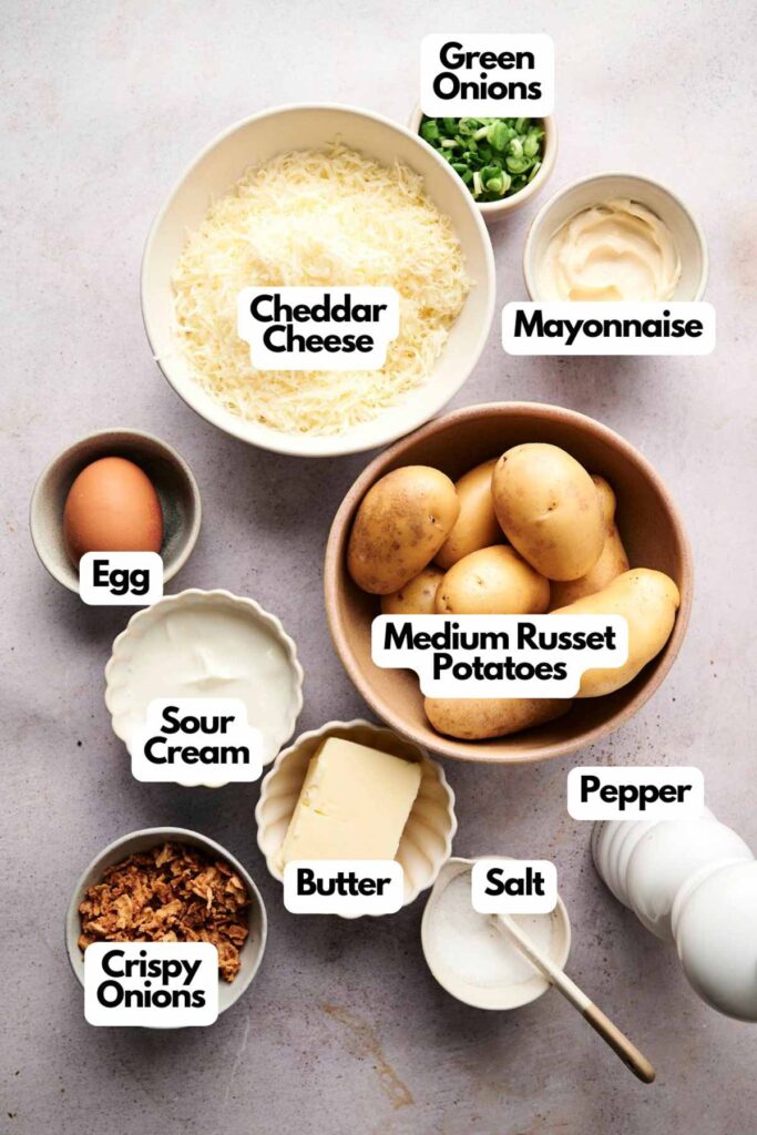 Ingredients for twice baked potatoes are arranged on a table, including cheddar cheese, mayonnaise, green onions, medium russet potatoes, pepper, salt, butter, sour cream, crispy onions, and an egg.