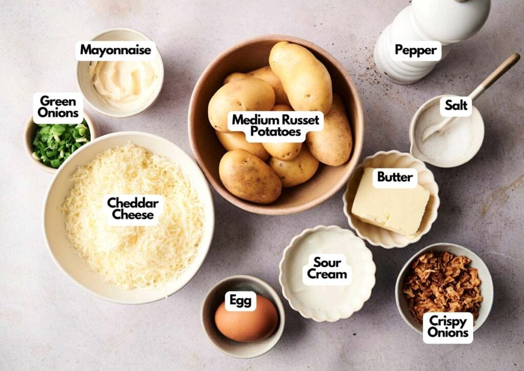 Top-down view of labeled ingredients for twice baked potatoes: medium russet potatoes, cheddar cheese, green onions, mayonnaise, egg, sour cream, butter, crispy onions, salt, and pepper.