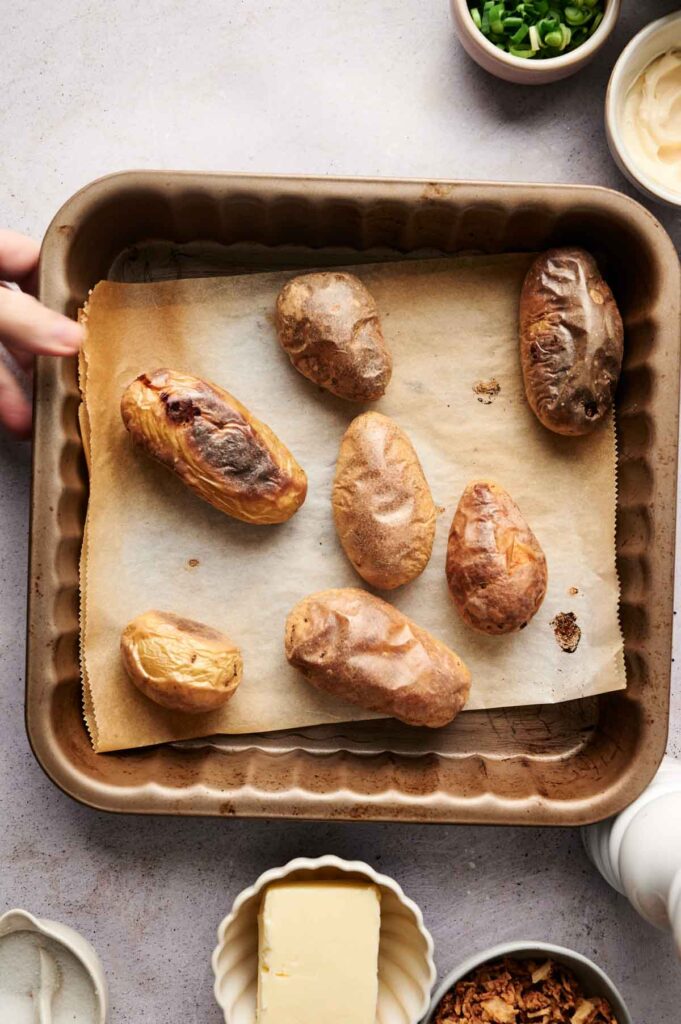 Twice baked potatoes rest on a parchment-lined tray, surrounded by ingredients like butter, green onions, mayonnaise, and other toppings.