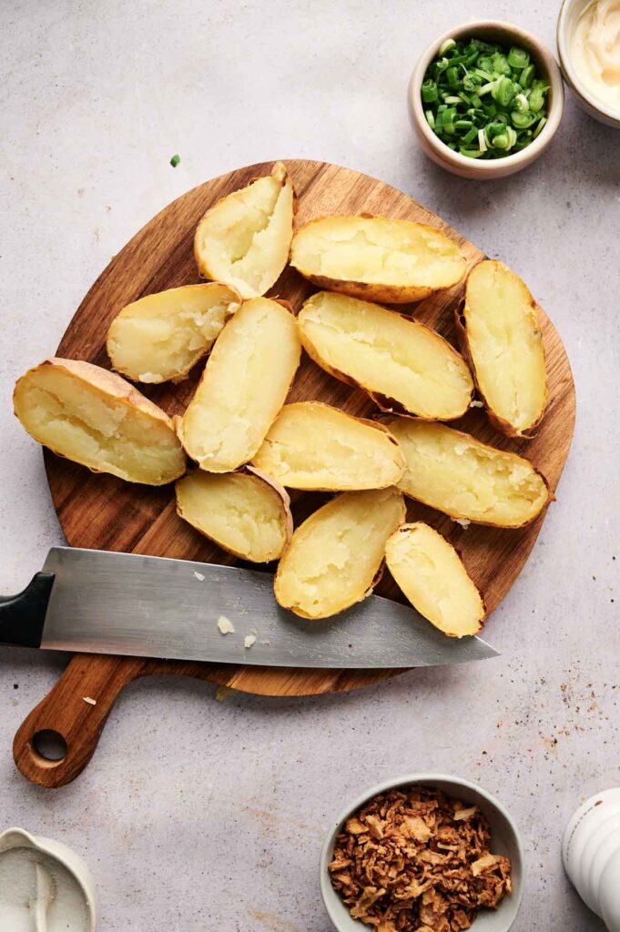 Sliced twice-baked potatoes rest on a wooden board with a knife, surrounded by bowls of green onions, fried onions, and sauce.