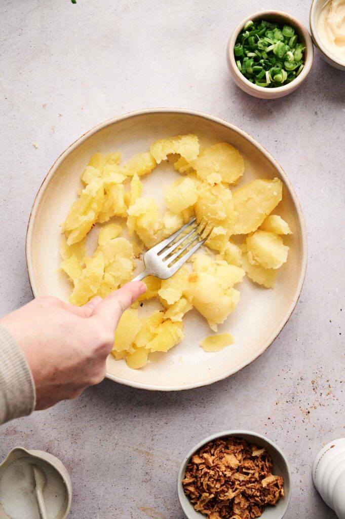 Hand mashing cooked potatoes with a fork in a bowl, reminiscent of twice baked potatoes, surrounded by bowls of green onions, crispy onions, and a creamy ingredient on a light surface.