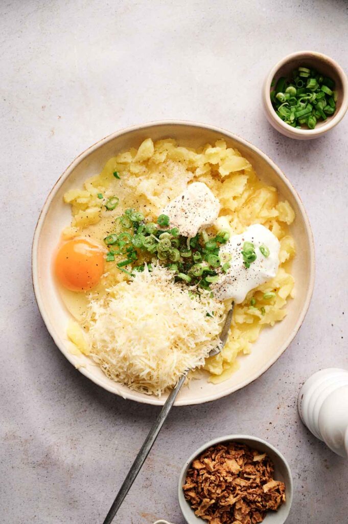 Bowl with mashed potatoes reminiscent of twice-baked potatoes, topped with a raw egg, grated cheese, green onions, and sour cream. Two small bowls on the side contain more green onions and crispy fried onions.
