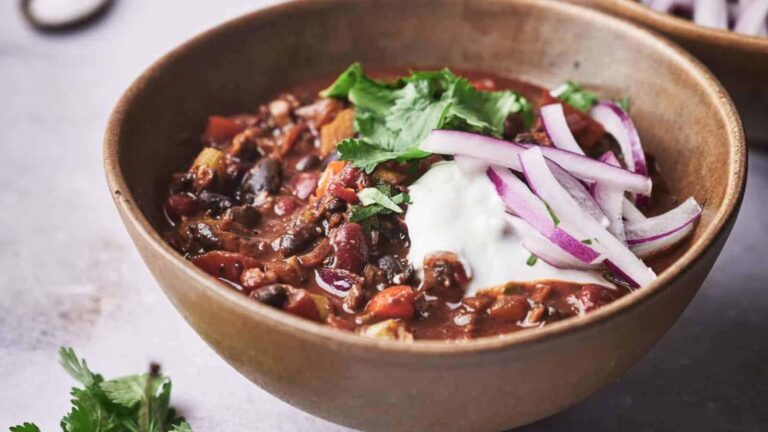 A bowl of chili topped with sliced red onions, a dollop of sour cream, and fresh cilantro, placed on a gray surface.