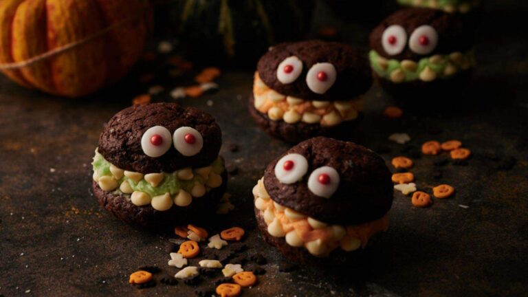 Chocolate cookies with candy eyes and colorful frosting resemble monster faces, surrounded by small festive sprinkles on a dark surface.