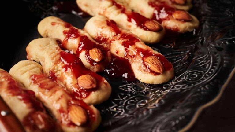 Cookies shaped like fingers with almonds as nails and red jam as decoration, placed on a dark patterned plate.