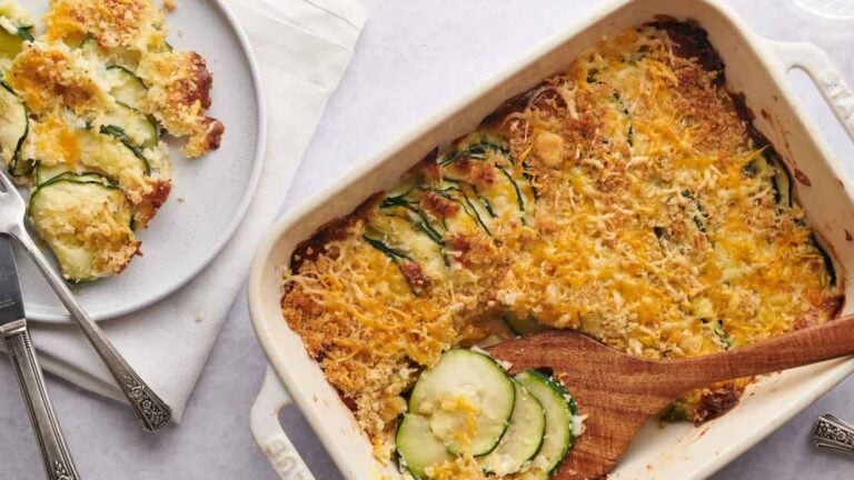 A baked zucchini casserole topped with melted cheese and breadcrumbs in a white dish. A portion is served on a white plate with a fork beside it.
