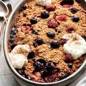 Oval dish with mixed berry crumble topped with whipped cream dollops. Spoons and bowl in background on a textured surface.