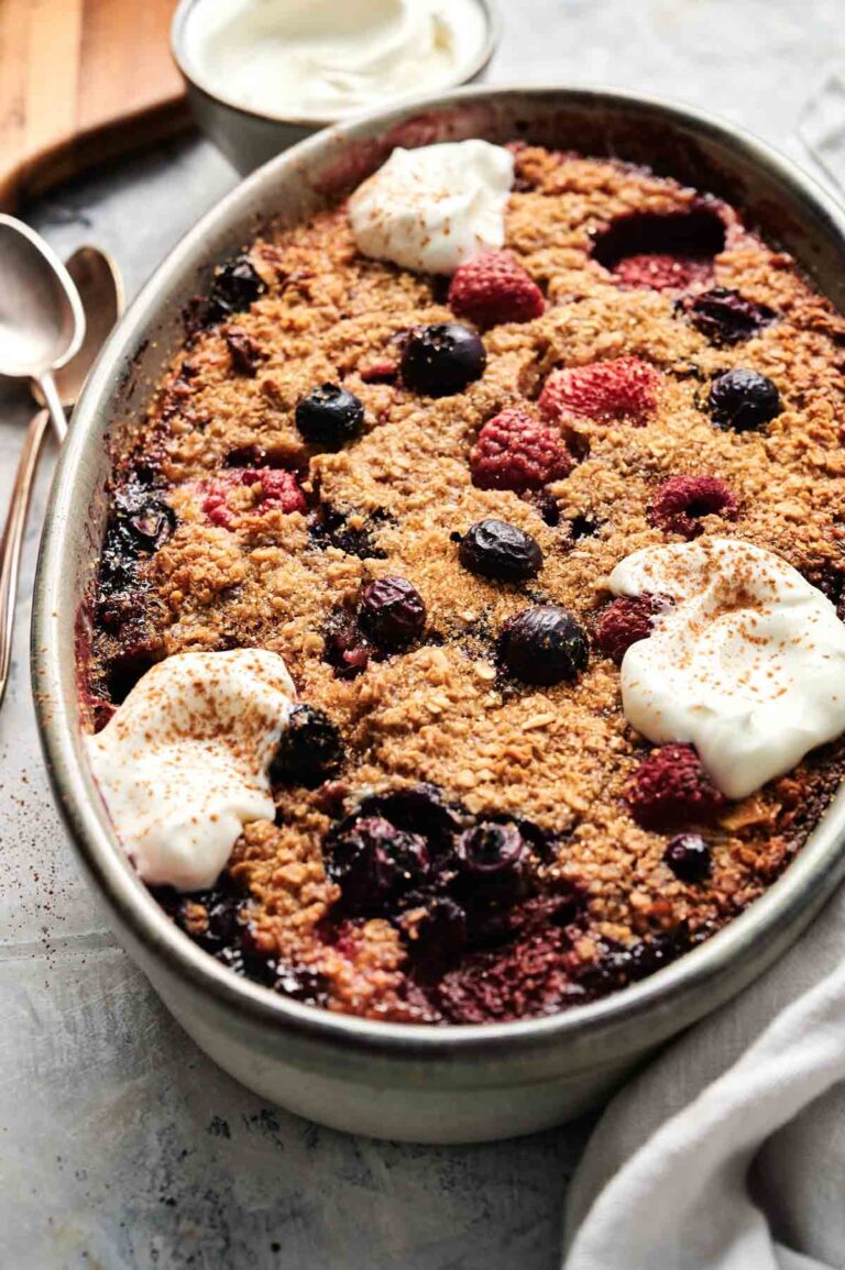 Oval dish with mixed berry crumble topped with whipped cream dollops. Spoons and bowl in background on a textured surface.