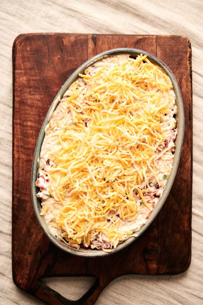 Oval dish on a wooden board, filled with a creamy Cattle Drive Casserole mixture topped with shredded cheese, ready for baking.
