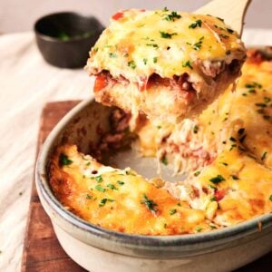 A serving of cheesy Cattle Drive Casserole is being lifted from a baking dish with a wooden spoon. The casserole is garnished with fresh herbs, promising a hearty and savory delight.