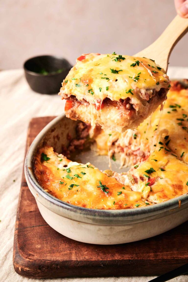 A serving of cheesy Cattle Drive Casserole is being lifted from a baking dish with a wooden spoon. The casserole is garnished with fresh herbs, promising a hearty and savory delight.
