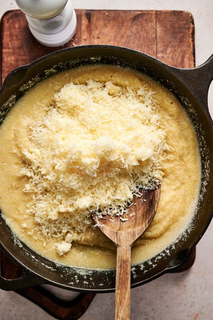 A cast iron skillet with creamy cheese grits topped with grated cheese, stirred with a wooden spoon on a wooden board.