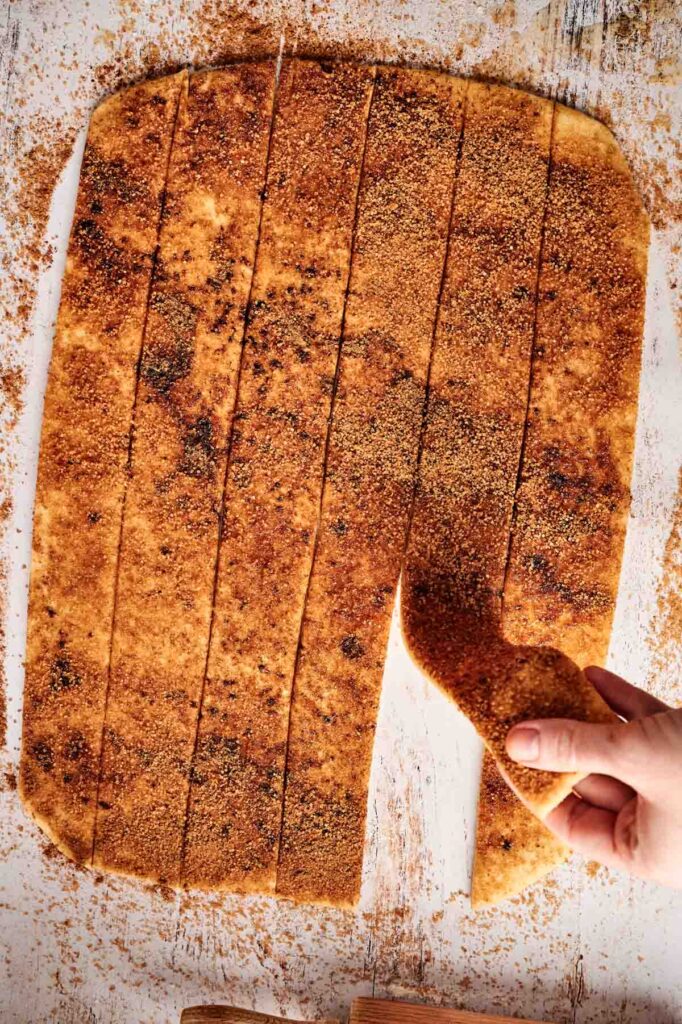 A hand peeling a strip from a cinnamon roll cake, coated in sugar and cinnamon, on a floured surface.