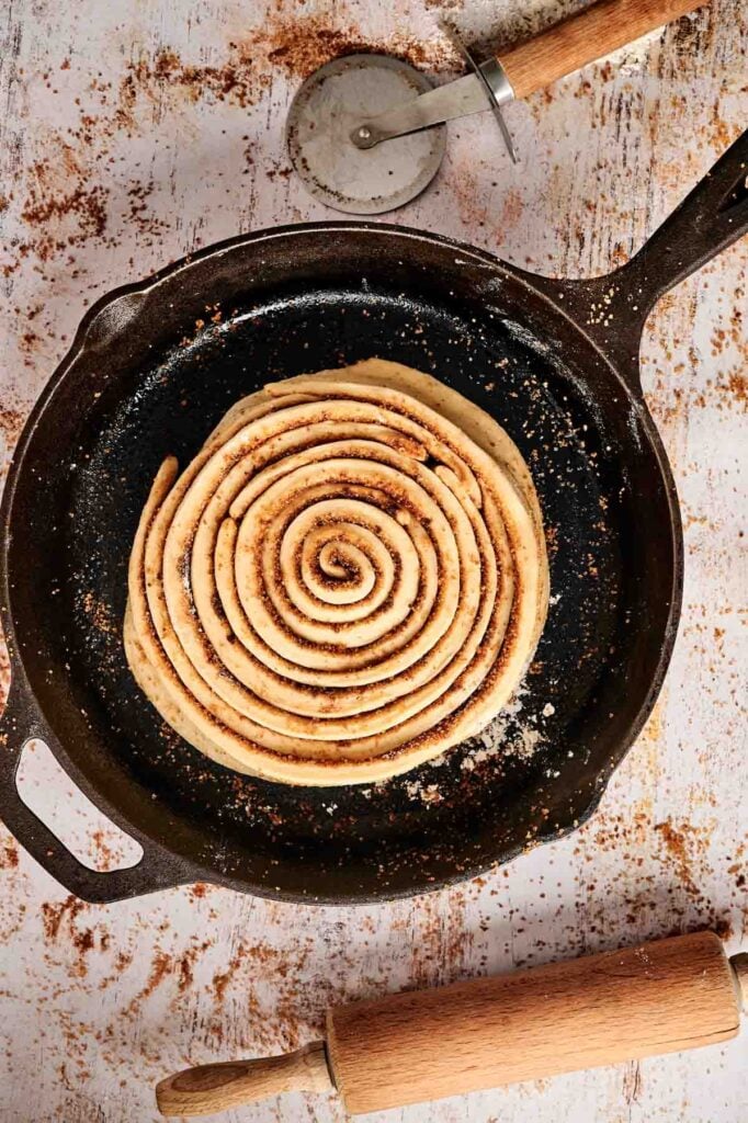 A rustic white surface showcases a cinnamon roll cake spiraled in a cast iron pan, with a pizza cutter and rolling pin nearby.