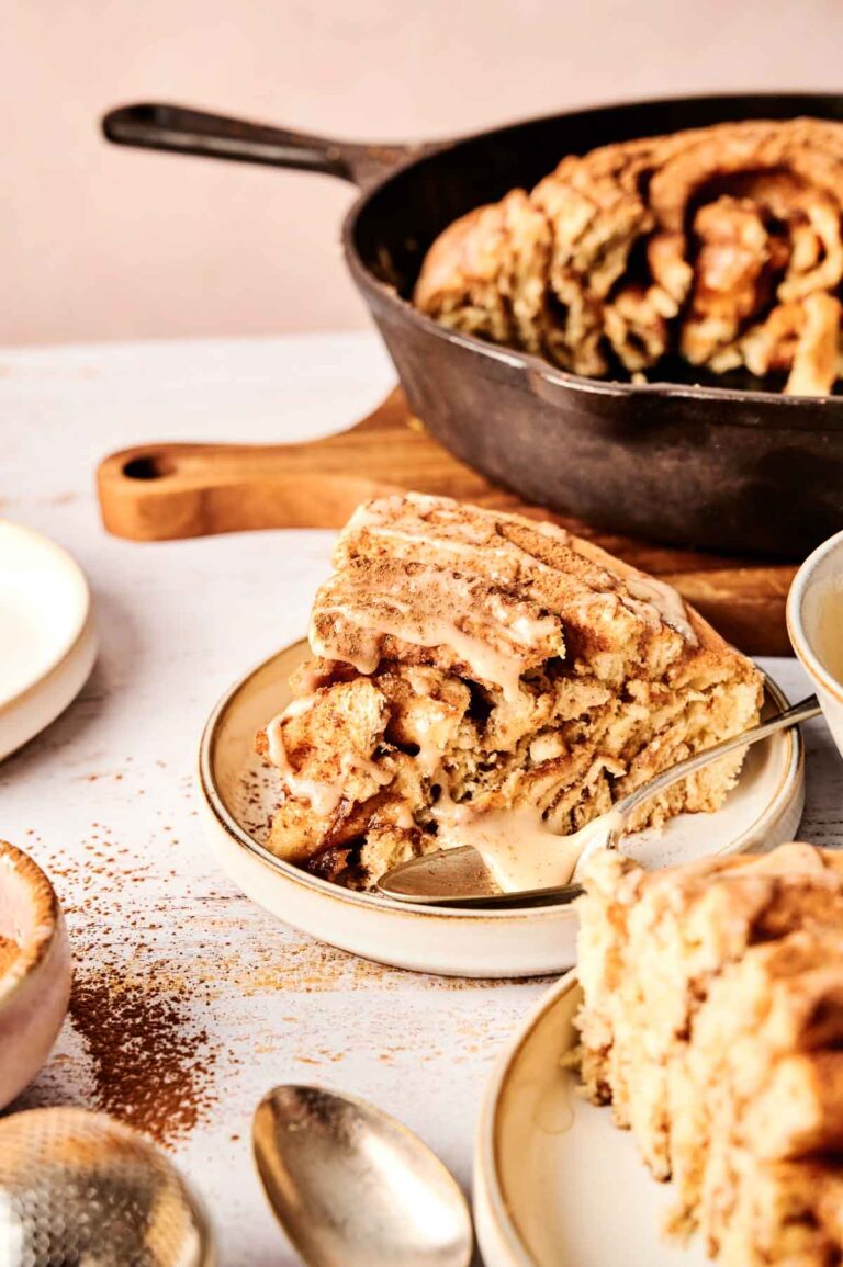 A slice of delicious cinnamon roll cake with icing rests on a plate, accompanied by a fork. In the background, a cast iron pan holds the remaining cake. Spoons are scattered on the table, inviting you to indulge in this delightful treat.
