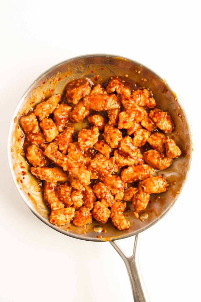 A pan filled with crispy General Tso’s Chicken pieces, each coated in a sticky, reddish-brown sauce, set against a pristine white backdrop.