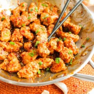 Stir-fried chicken pieces, reminiscent of General Tso’s Chicken, are coated in a glistening spicy sauce with sesame seeds, garnished with chopped green herbs and served with chopsticks.
