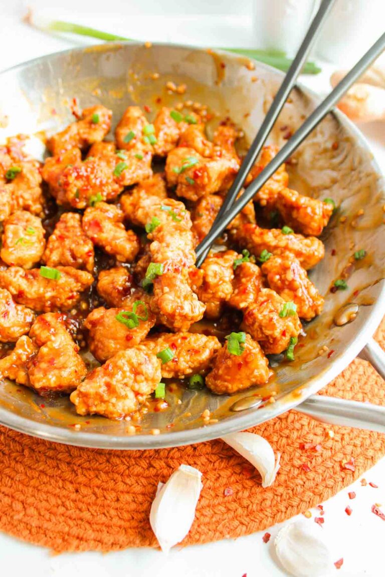 Stir-fried chicken pieces, reminiscent of General Tso’s Chicken, are coated in a glistening spicy sauce with sesame seeds, garnished with chopped green herbs and served with chopsticks.