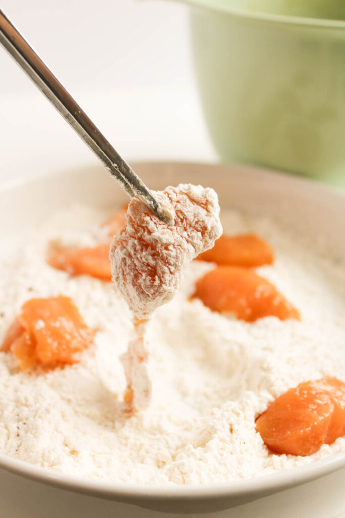 A chicken piece, reminiscent of General Tso’s Chicken, is coated in flour and spices, held by tongs over a white bowl filled with more seasoned pieces.