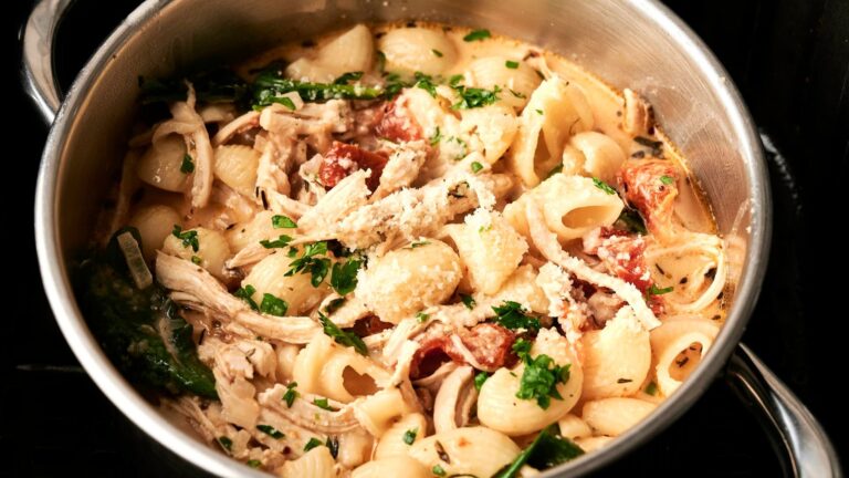 A pot of pasta with shell-shaped noodles, shredded chicken, spinach, cherry tomatoes, and a creamy sauce, garnished with grated cheese and parsley.