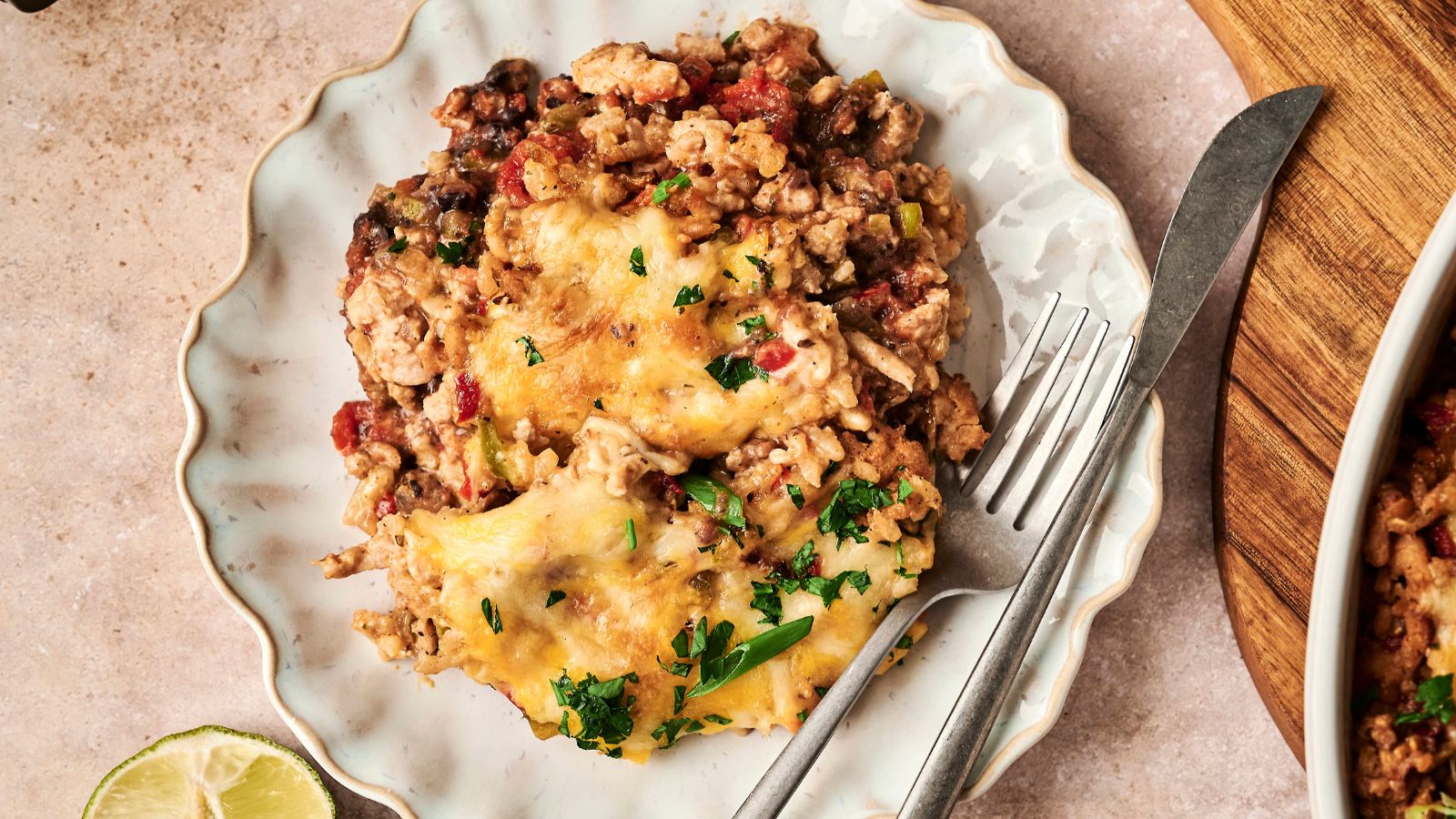 A plate with a serving of cheesy casserole containing ground meat, beans, and chopped herbs, accompanied by a fork and knife. A lime wedge is partially visible on the side.