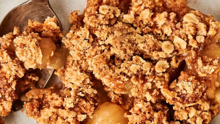 Close-up of apple crumble with a metal spoon, showing a golden-brown topping made of oats and crumbled pieces over baked apple slices.