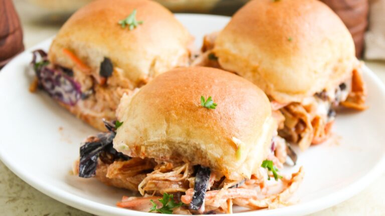 Three pulled pork sliders with coleslaw on a white plate, topped with a sprig of parsley.